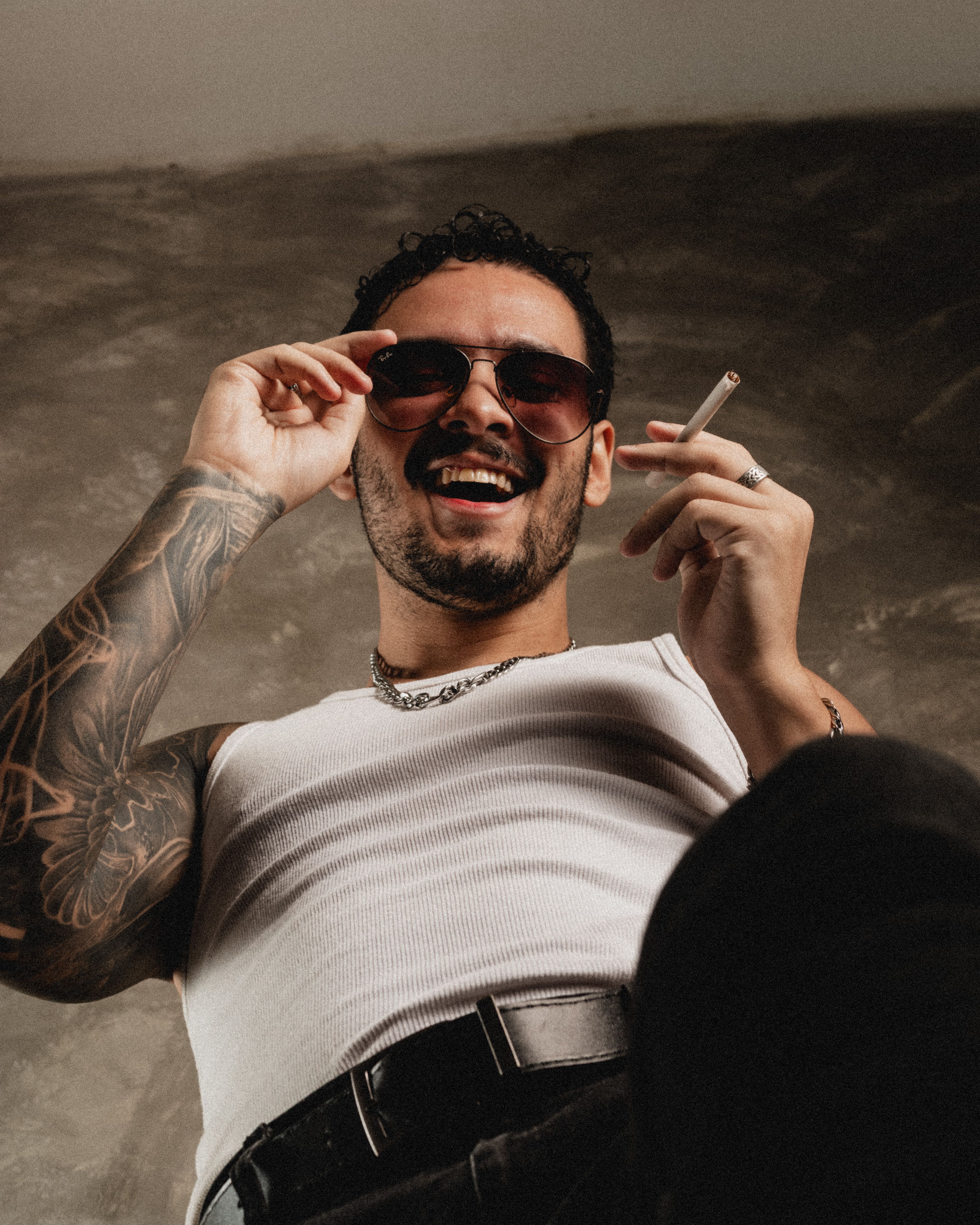 Upward angle shot of a stylish man with tattoos and sunglasses enjoying a cigarette indoors.