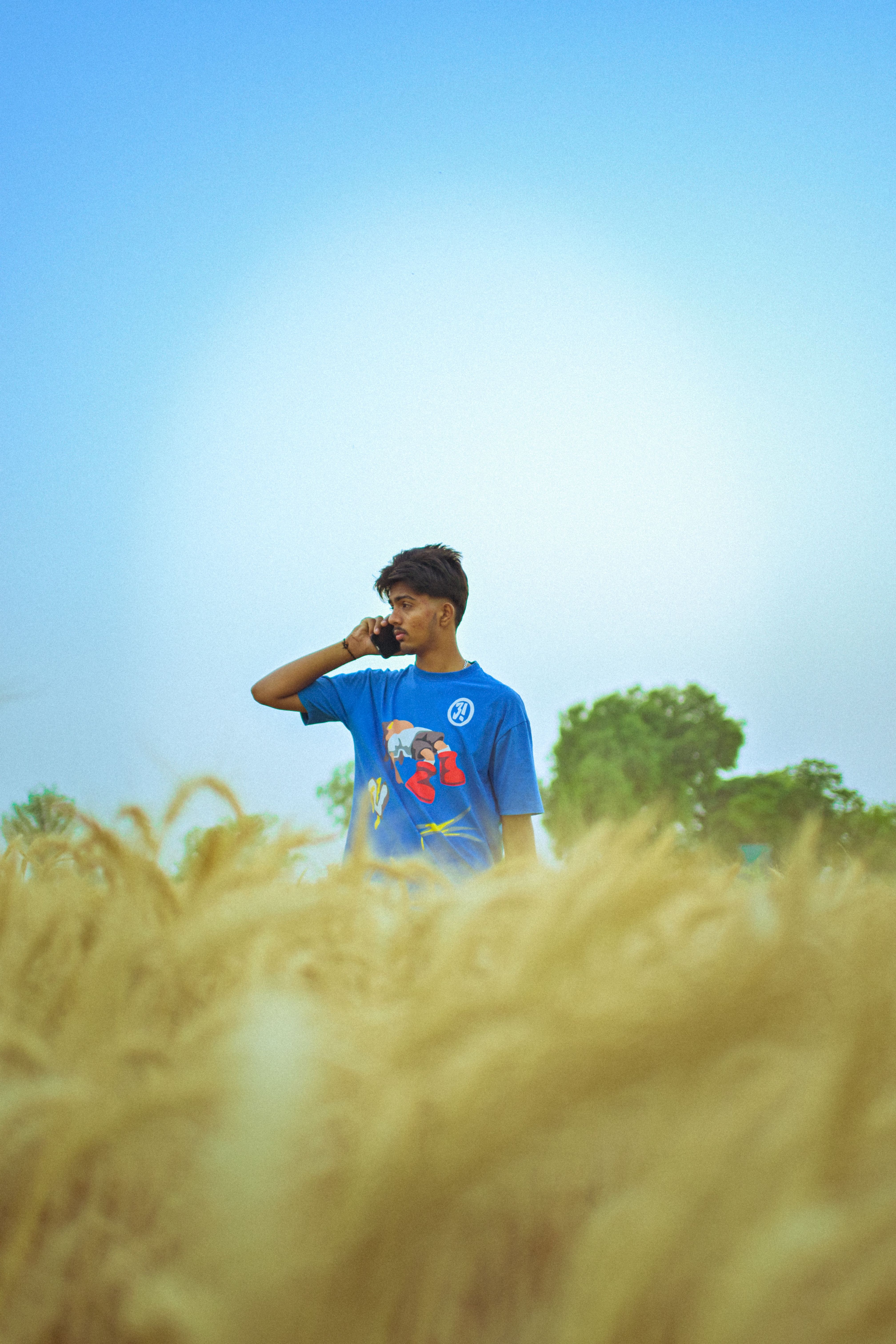 A young man in a blue t-shirt talking on the phone, standing in a lush wheat field under a clear sky.