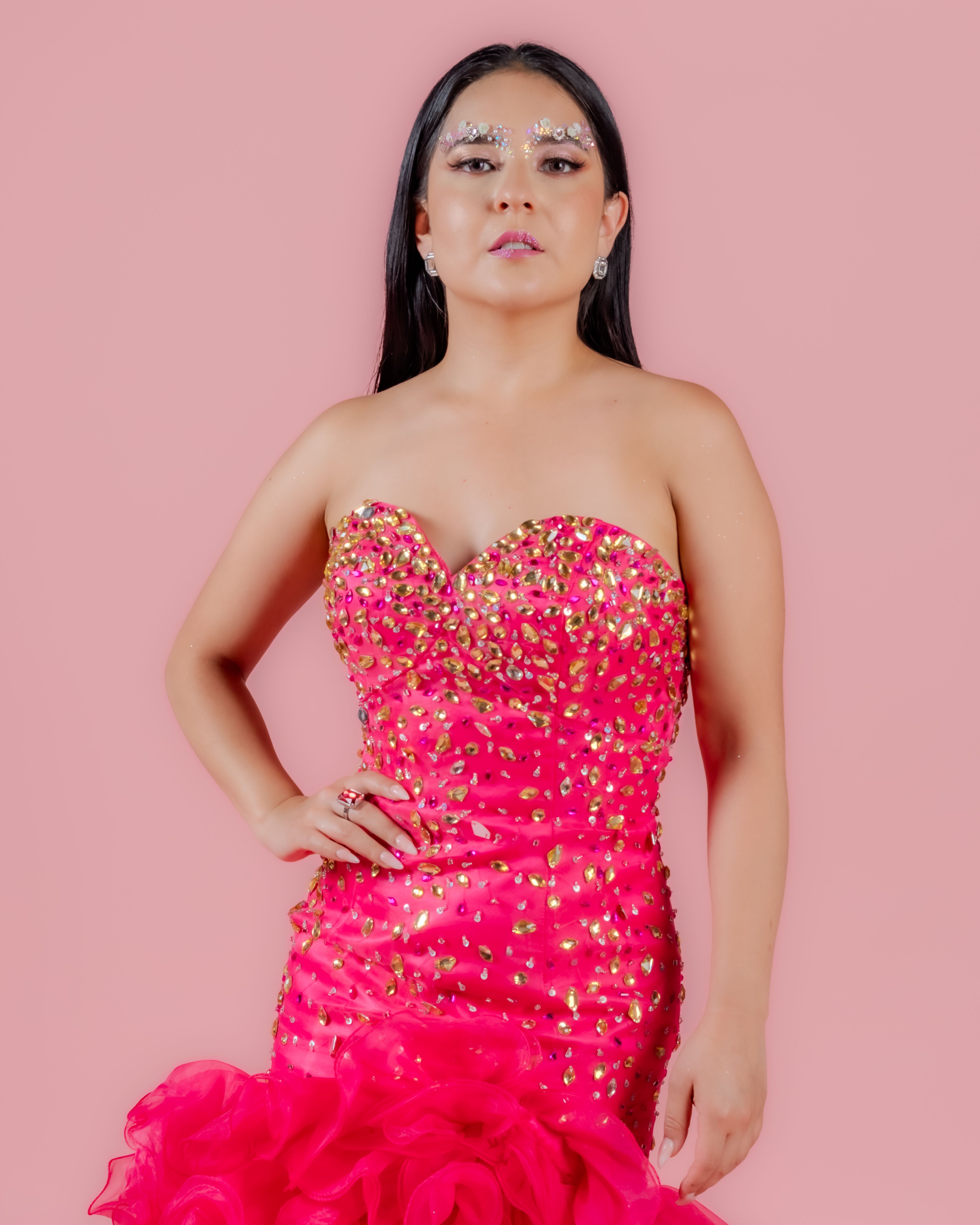 A confident woman poses elegantly in a sparkling pink gown against a pastel background.