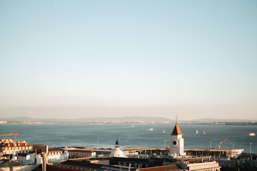 A scenic view of a coastal town with a clock tower overlooking the calm ocean and sailboats.