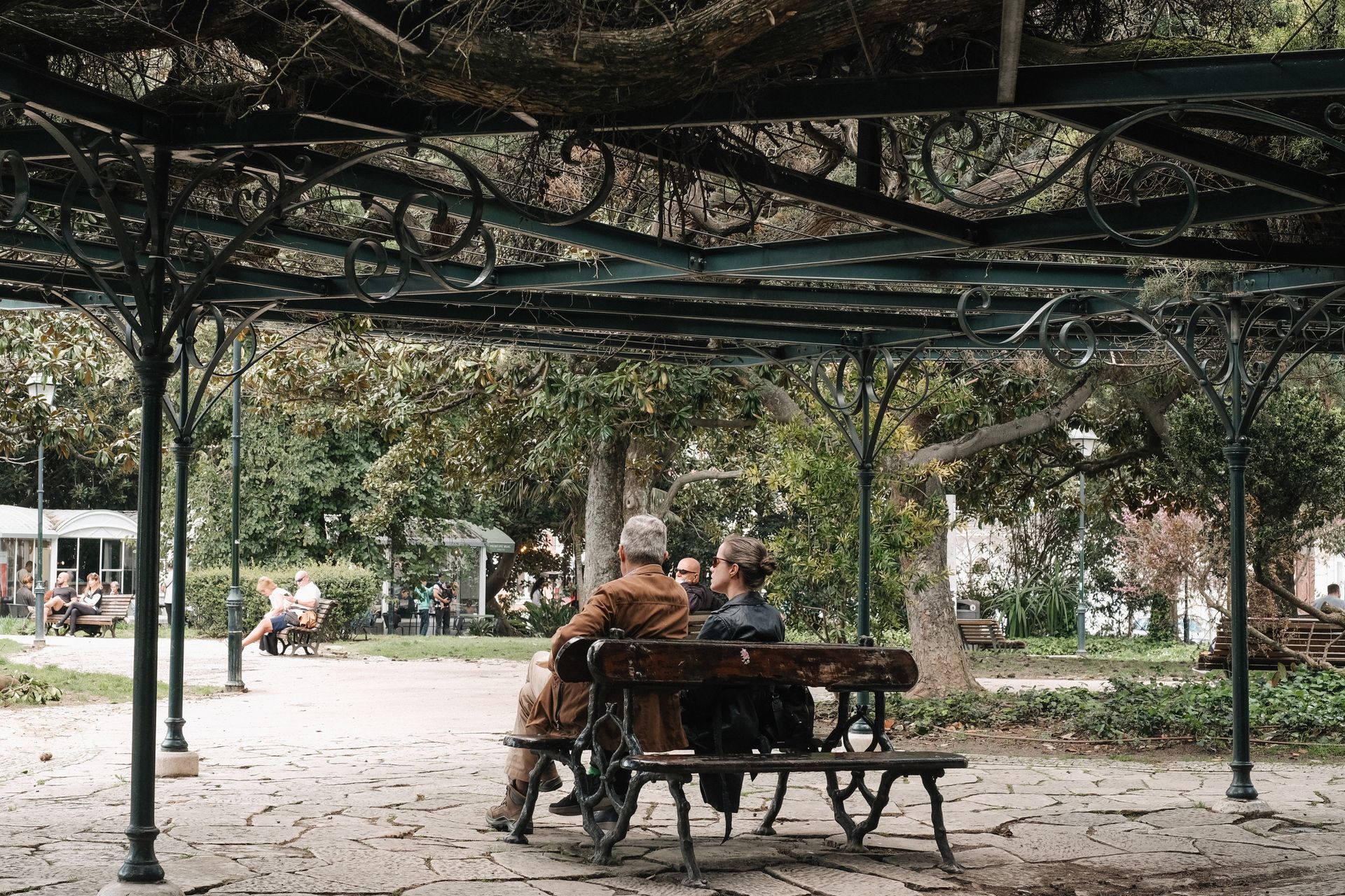 A peaceful scene of two people conversing under a decorative gazebo in a lush outdoor park.