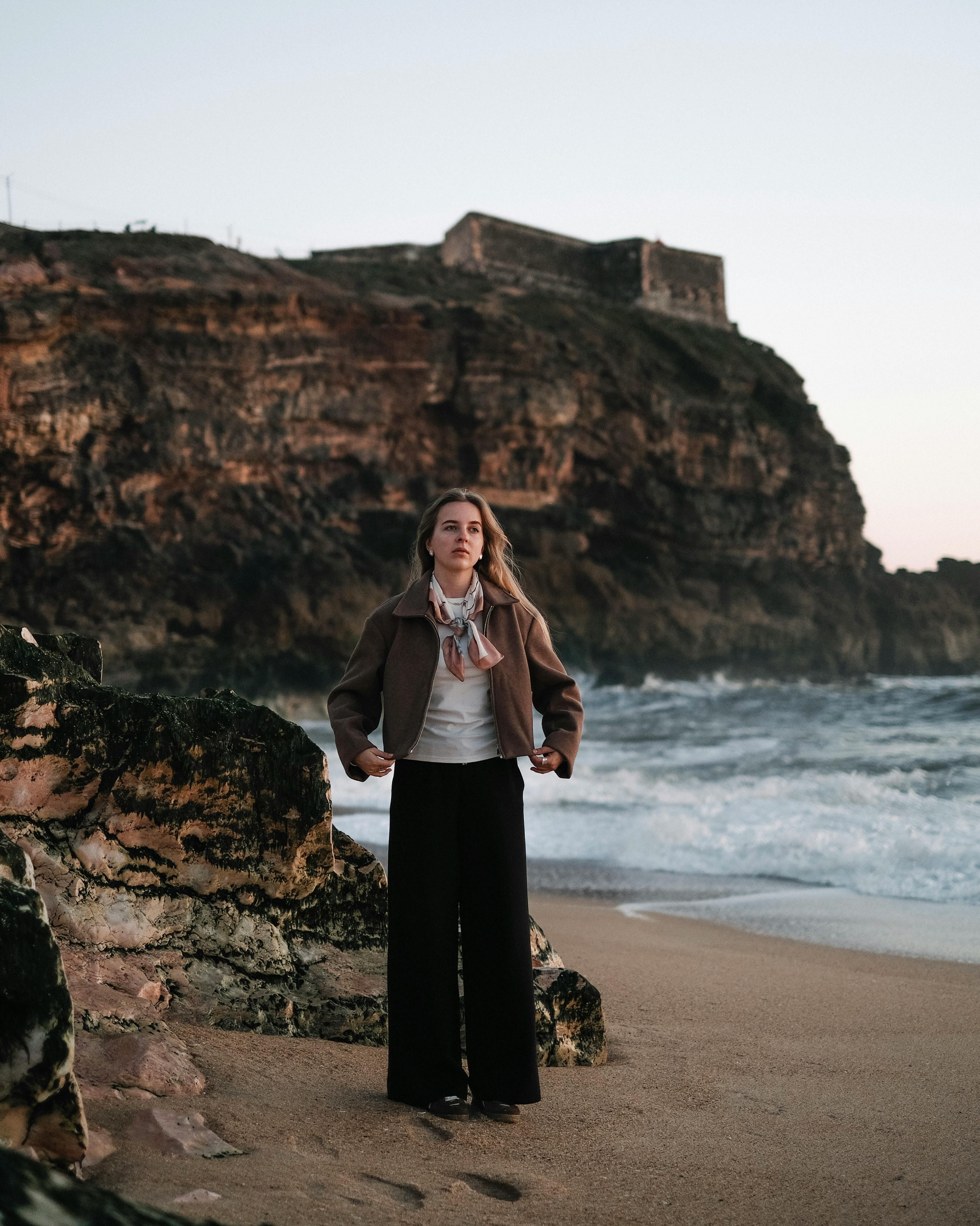 Young woman standing by rugged cliffs with ocean waves at sunset, exuding calm and contemplation.