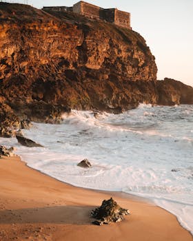 Dramatic cliff with fortress overlooking rugged ocean waves at sunset, creating a scenic coastal view.