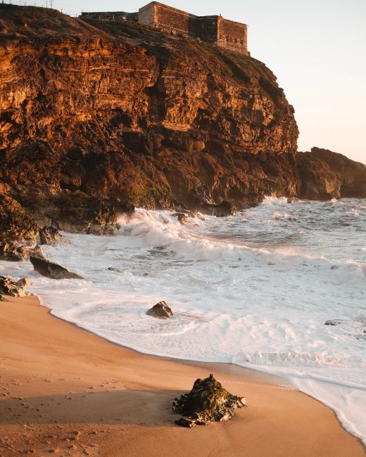 Dramatic cliff with fortress overlooking rugged ocean waves at sunset, creating a scenic coastal view.