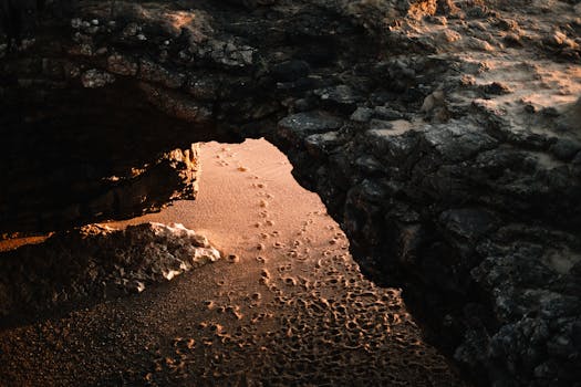 An illuminated coastal cave entrance with footprints in the sand at sunset, creating a dramatic scene.