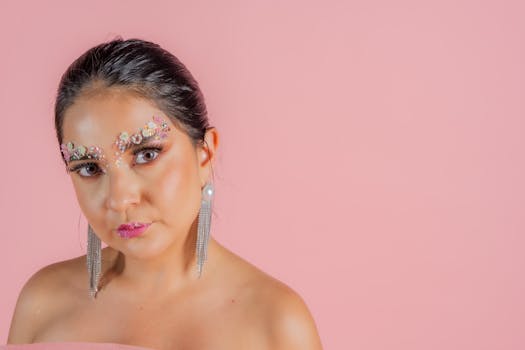 Stylish artistic portrait of a woman with creative makeup and elegant earrings against a pink background.