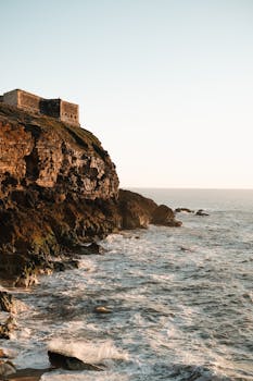A stunning view of a coastal fortress perched on a cliff at sunrise, overlooking the ocean waves.