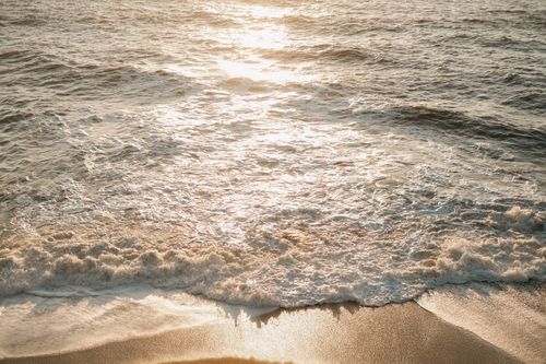 Free Sunset scene of ocean waves gently crashing on a sandy beach with golden sunlight reflections. Stock Photo
