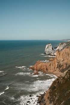 Majestic coastal cliffs rising above the turbulent ocean waves under a clear blue sky.