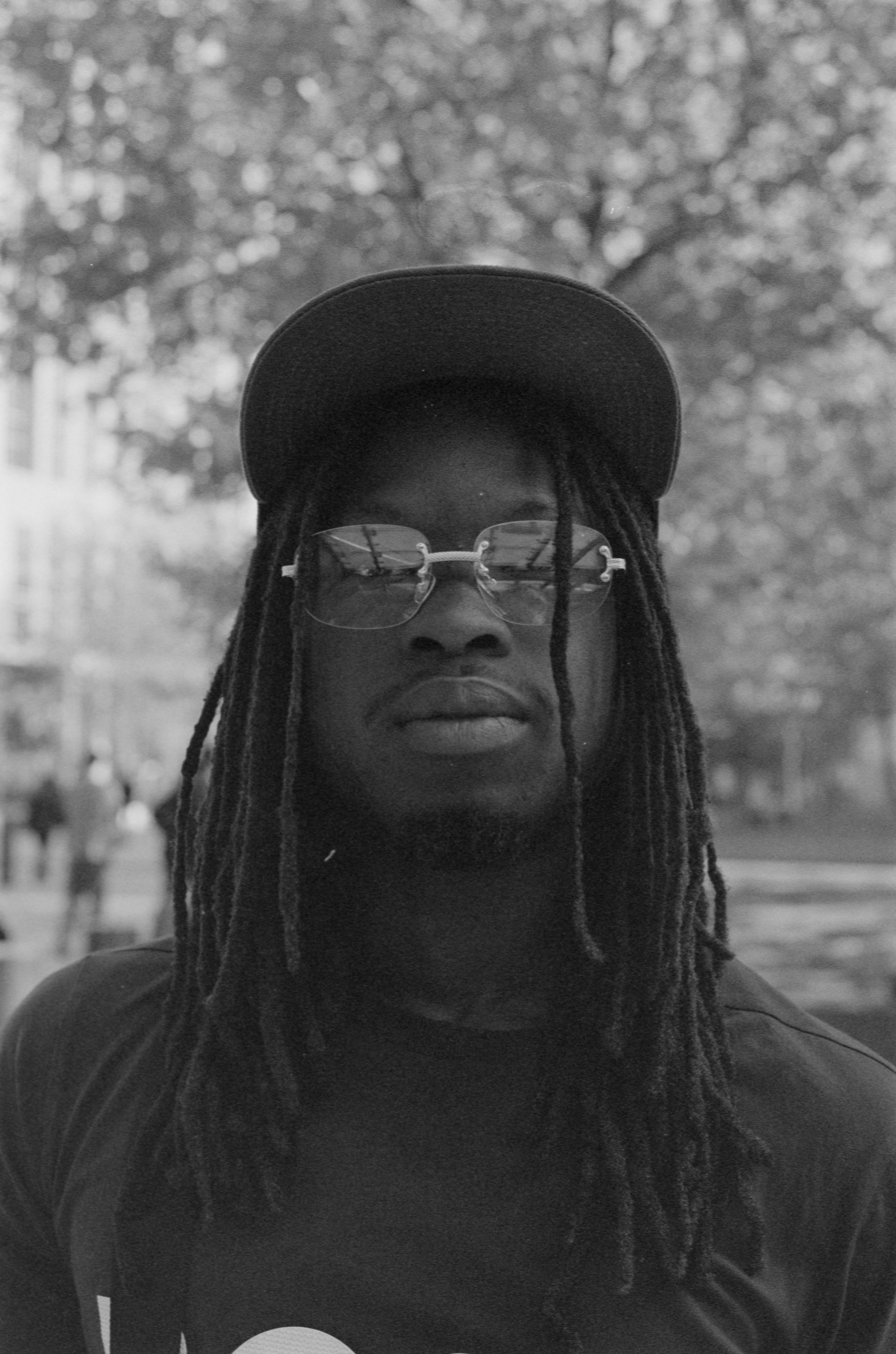 Black and white portrait of a young man wearing sunglasses and a cap outdoors.