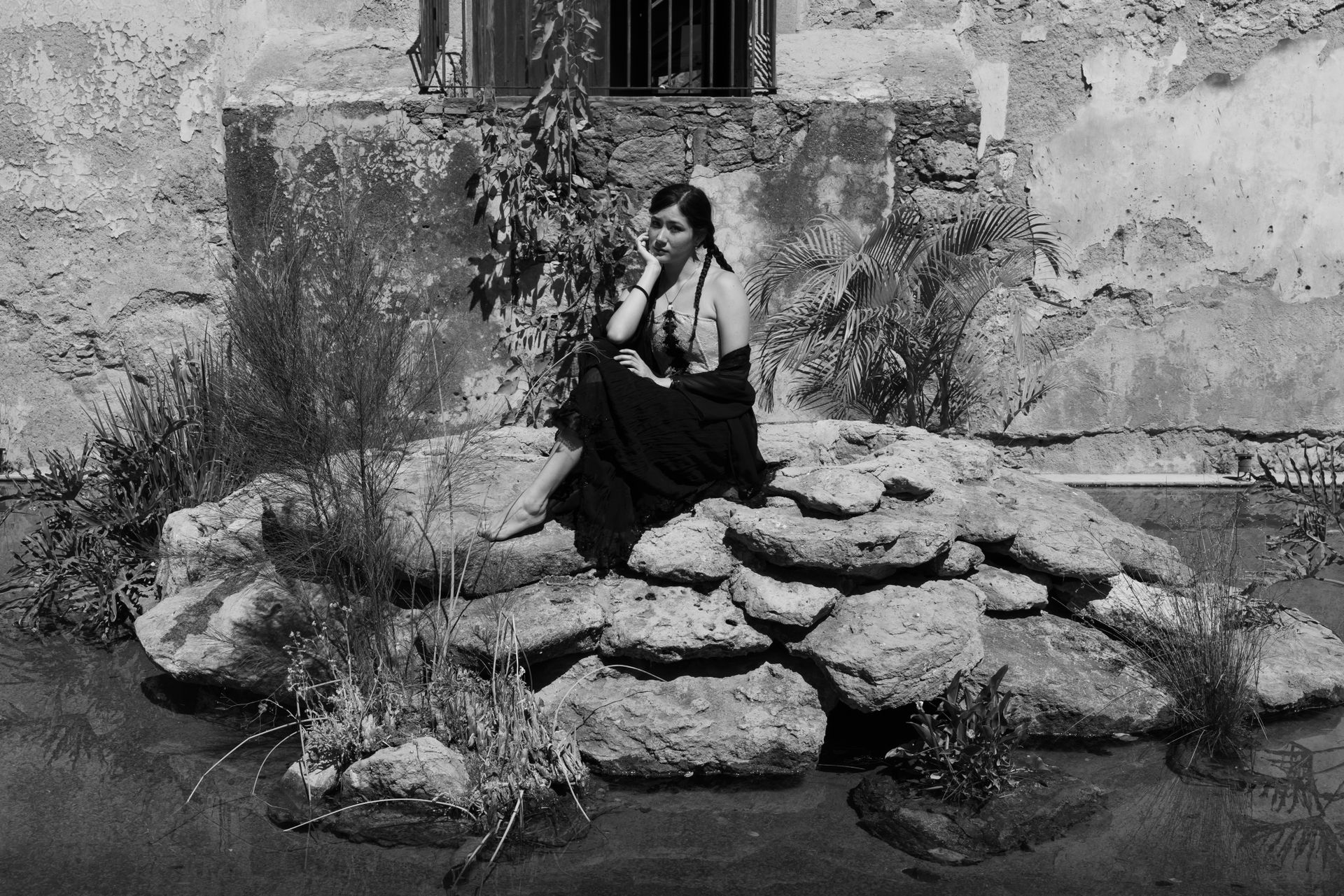 Elegant black and white portrait of a woman sitting pensively on rocks in a rustic garden setting.