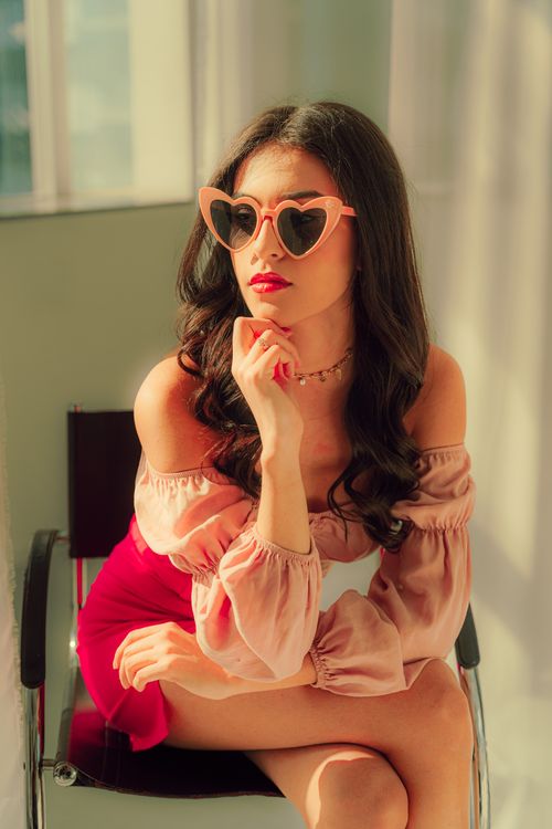 Free A fashionable woman wearing heart-shaped sunglasses and a stylish outfit sits elegantly in a sunlit room. Stock Photo