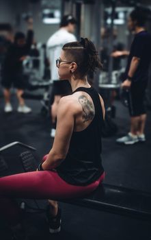 Athletic woman with owl tattoo resting on a bench in a gym, surrounded by people working out.