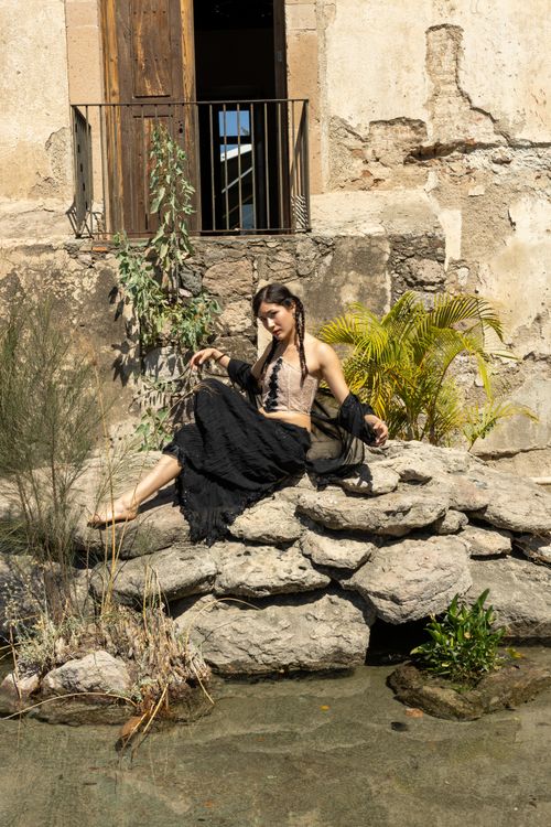 Free A young woman dressed in black and beige poses on rocks against a rustic wall outdoors. Stock Photo