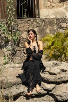 A woman in a black dress sitting on rocks against a rustic stone wall in sunlight.