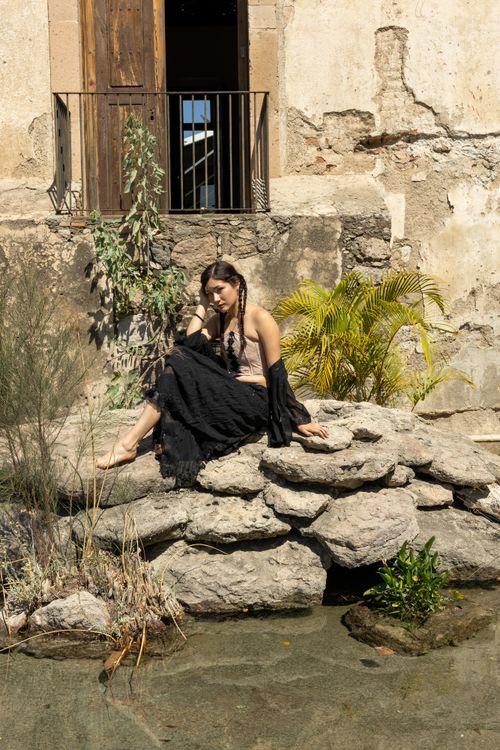 Free Woman in black dress sitting by a stone wall in a rustic outdoor setting under bright sunlight. Stock Photo