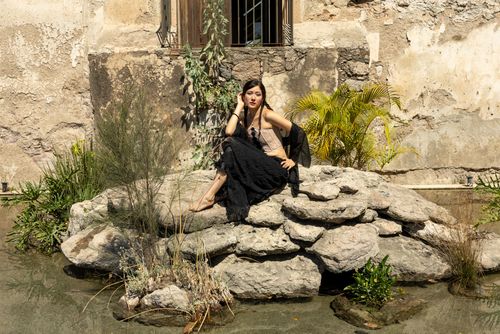 Free A woman in a dramatic pose sits on rocks against a rustic wall, surrounded by greenery. Stock Photo