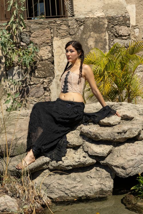 Free A woman in bohemian attire sits on rocks beside a historic stone wall and plants. Stock Photo