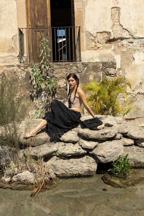 Free A woman in a stylish outfit sits gracefully on rocks by a rustic stone building, creating a serene outdoor scene. Stock Photo