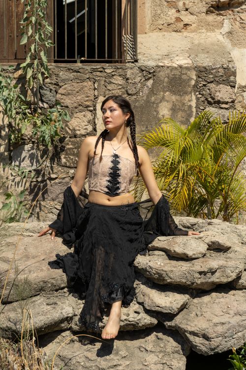 Free Woman in fashionable attire sits on stone wall with greenery background. Stock Photo