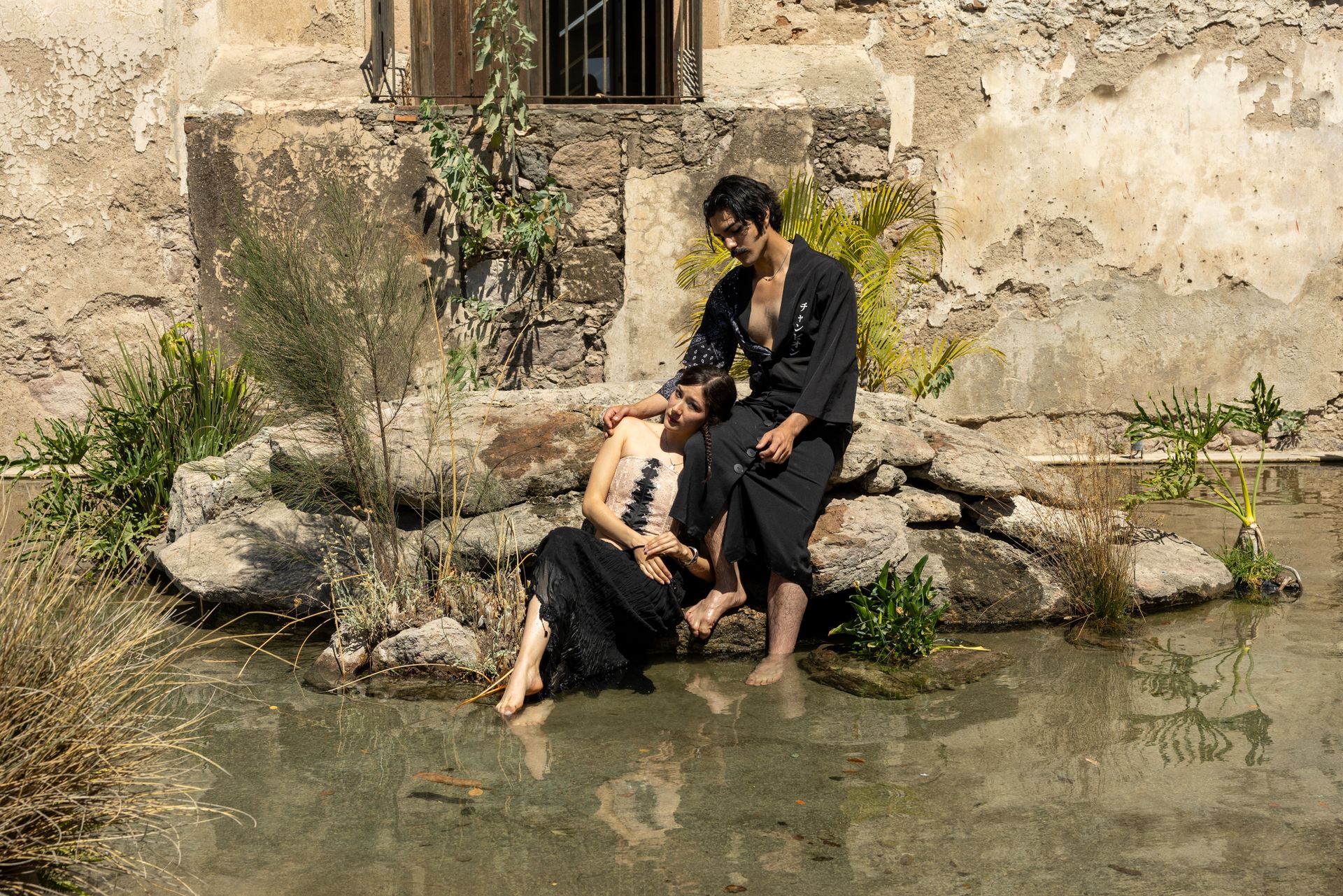 Couple in elegant attire relax by a water feature in a historic limestone courtyard.