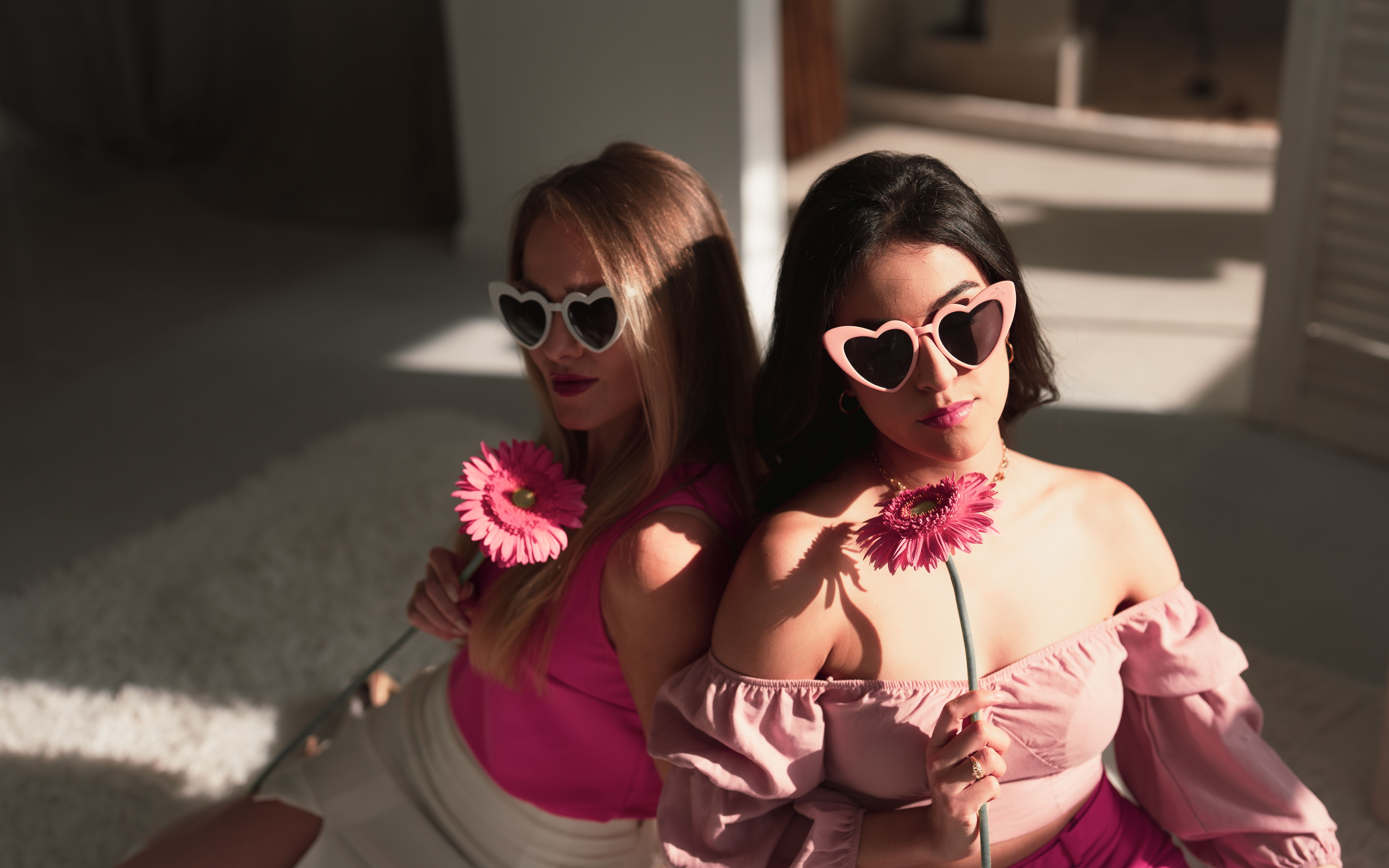 Two stylish women wearing pink heart sunglasses holding flowers. An indoor scene in soft light.