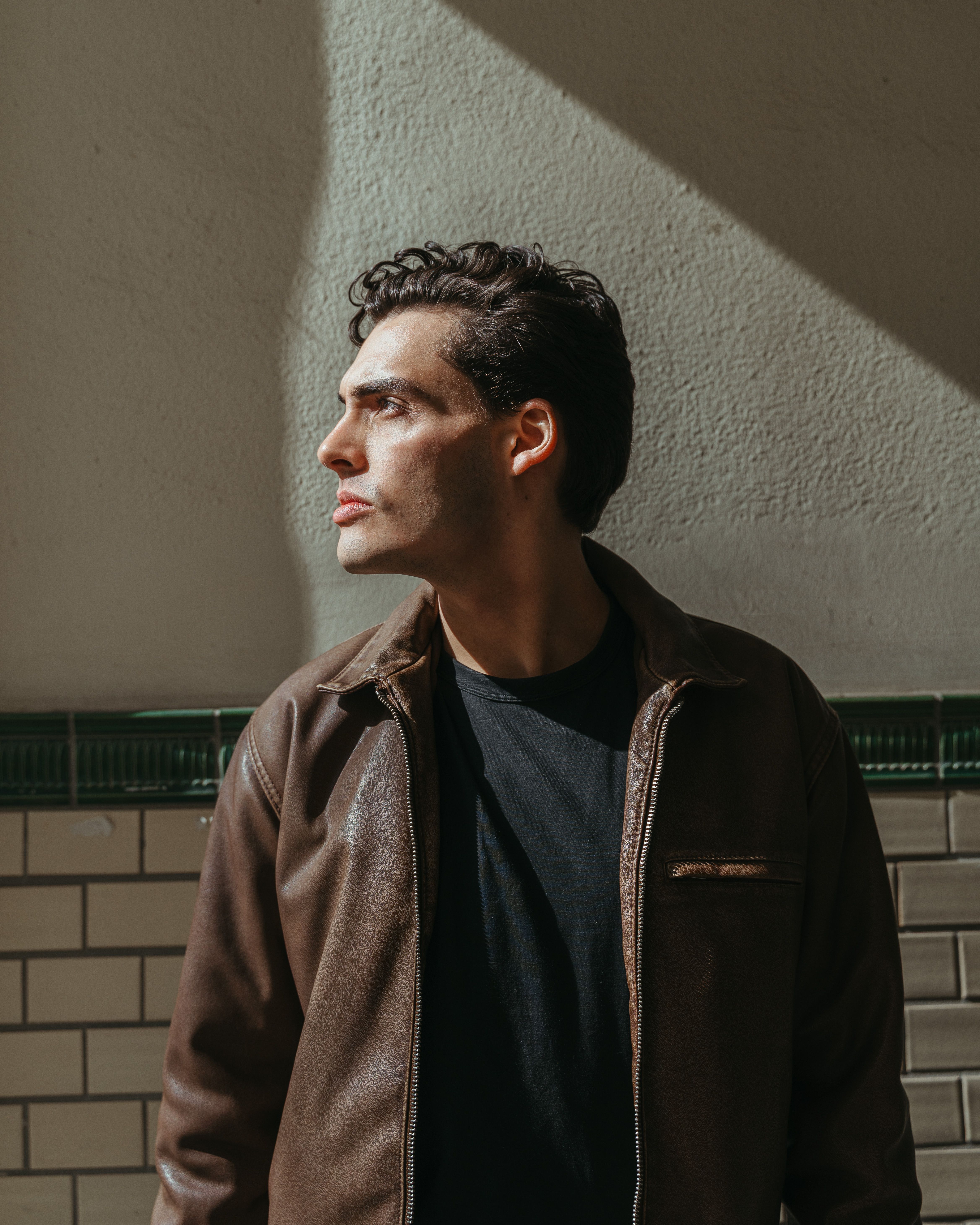 A side portrait of a young man in a leather jacket, basking in natural light indoors.