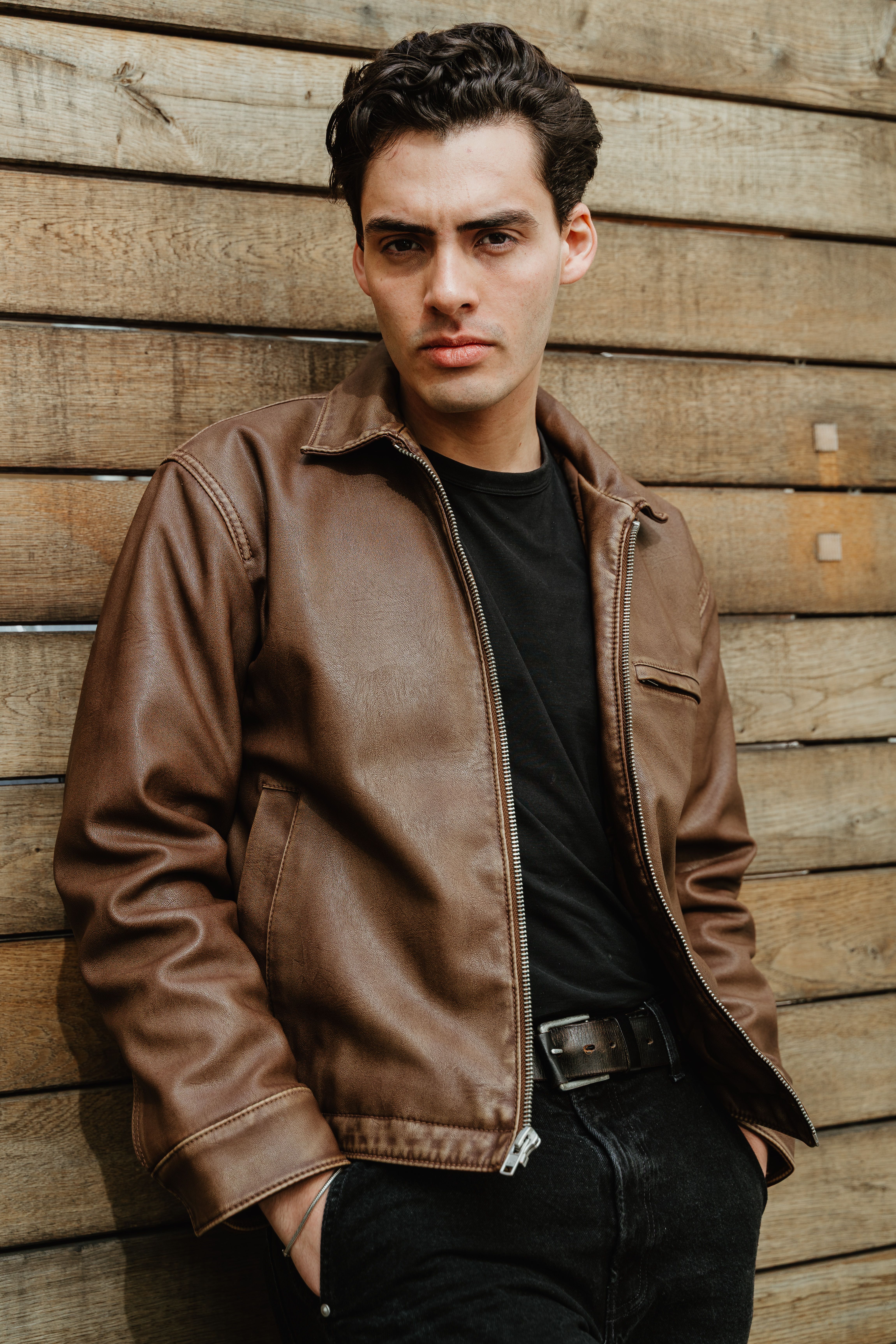 Outdoor portrait of a male model in Berlin wearing a brown leather jacket against a wooden backdrop.