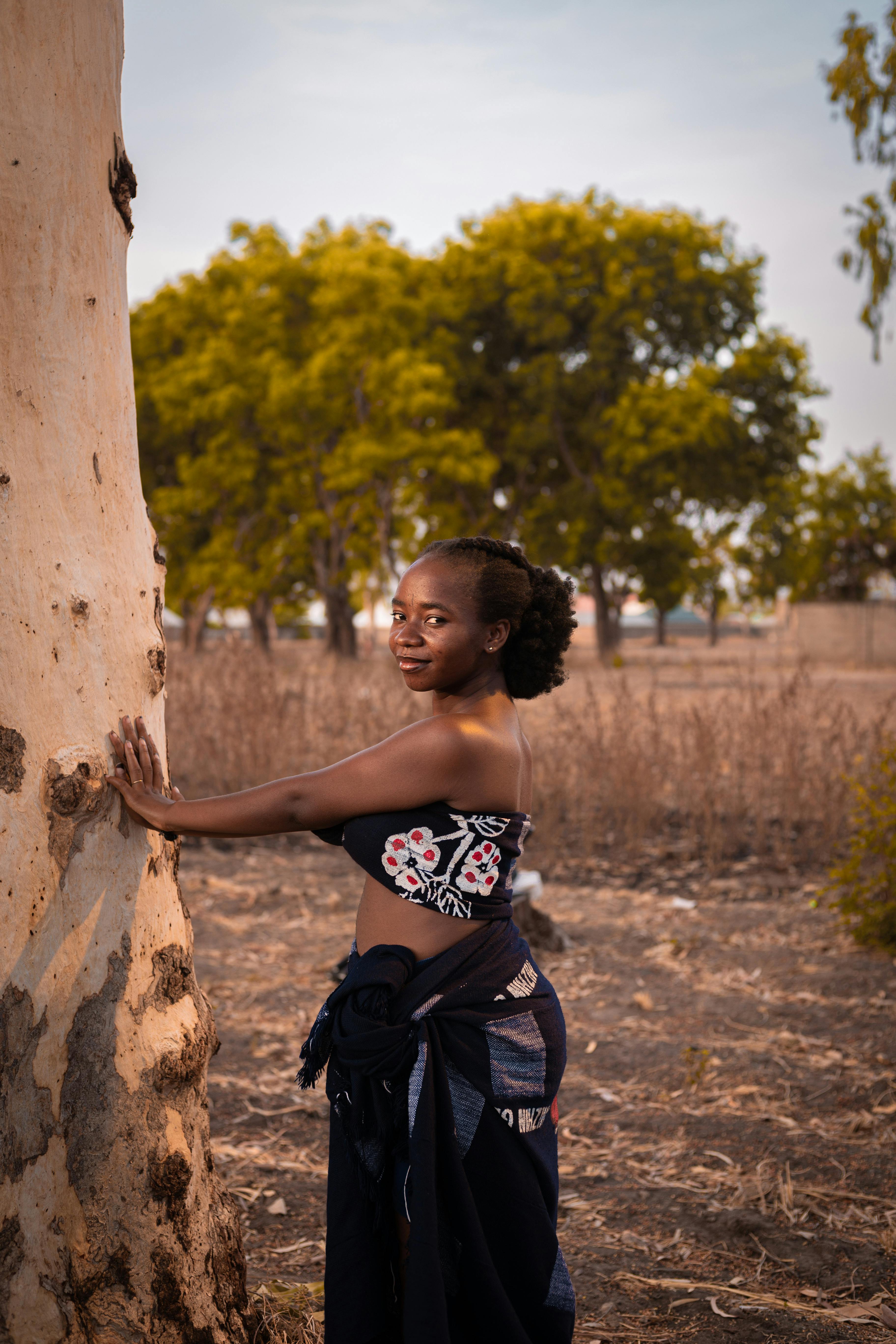 A smiling woman in traditional clothing poses outdoors by a tree under clear skies.
