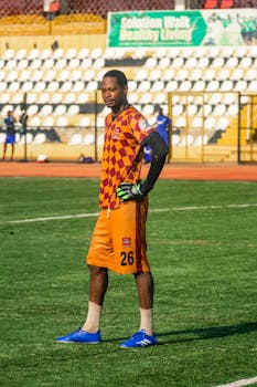 Male soccer player in orange and purple uniform standing on field during daytime.