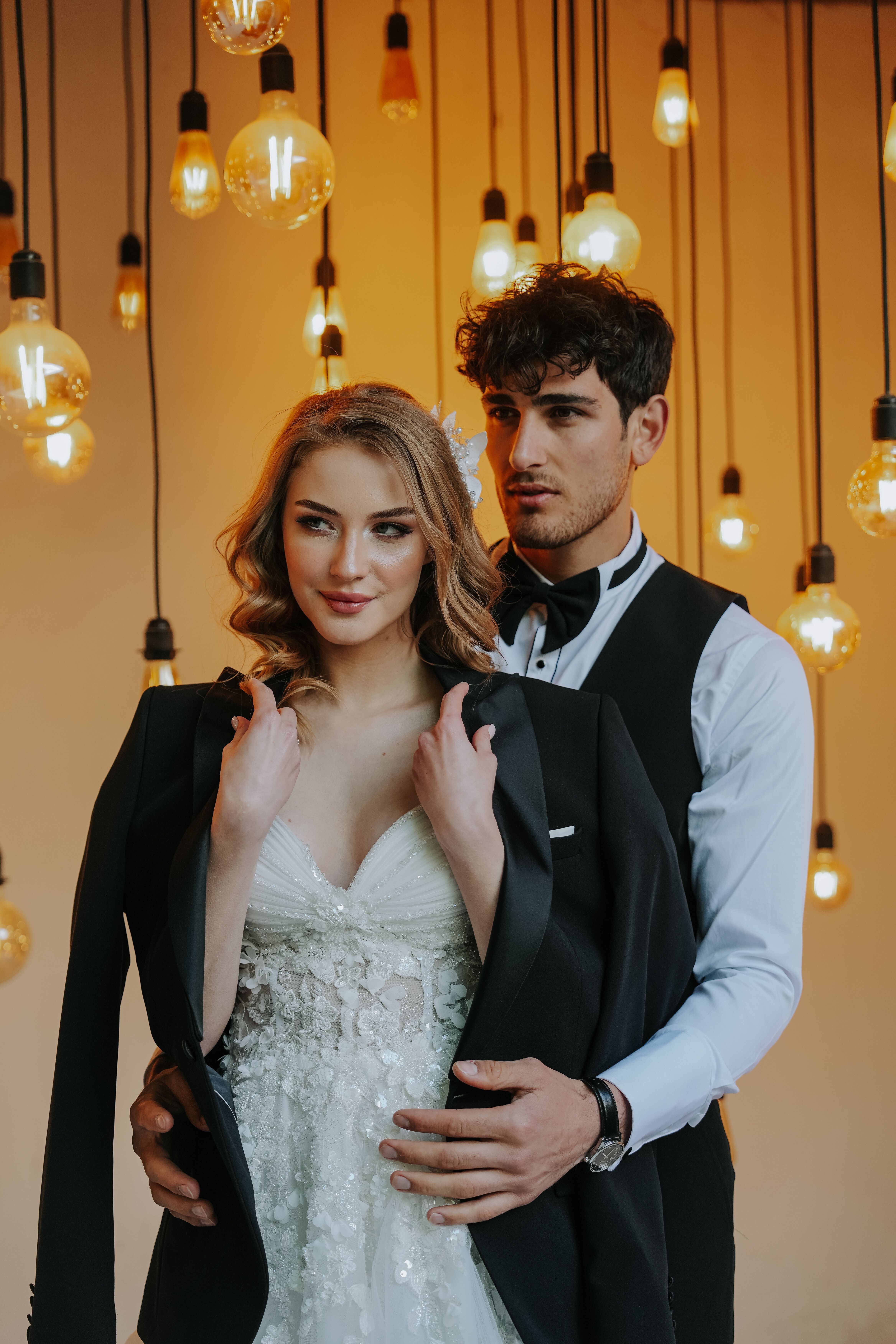 Stylish couple in formal attire surrounded by hanging lights, creating a romantic ambiance.