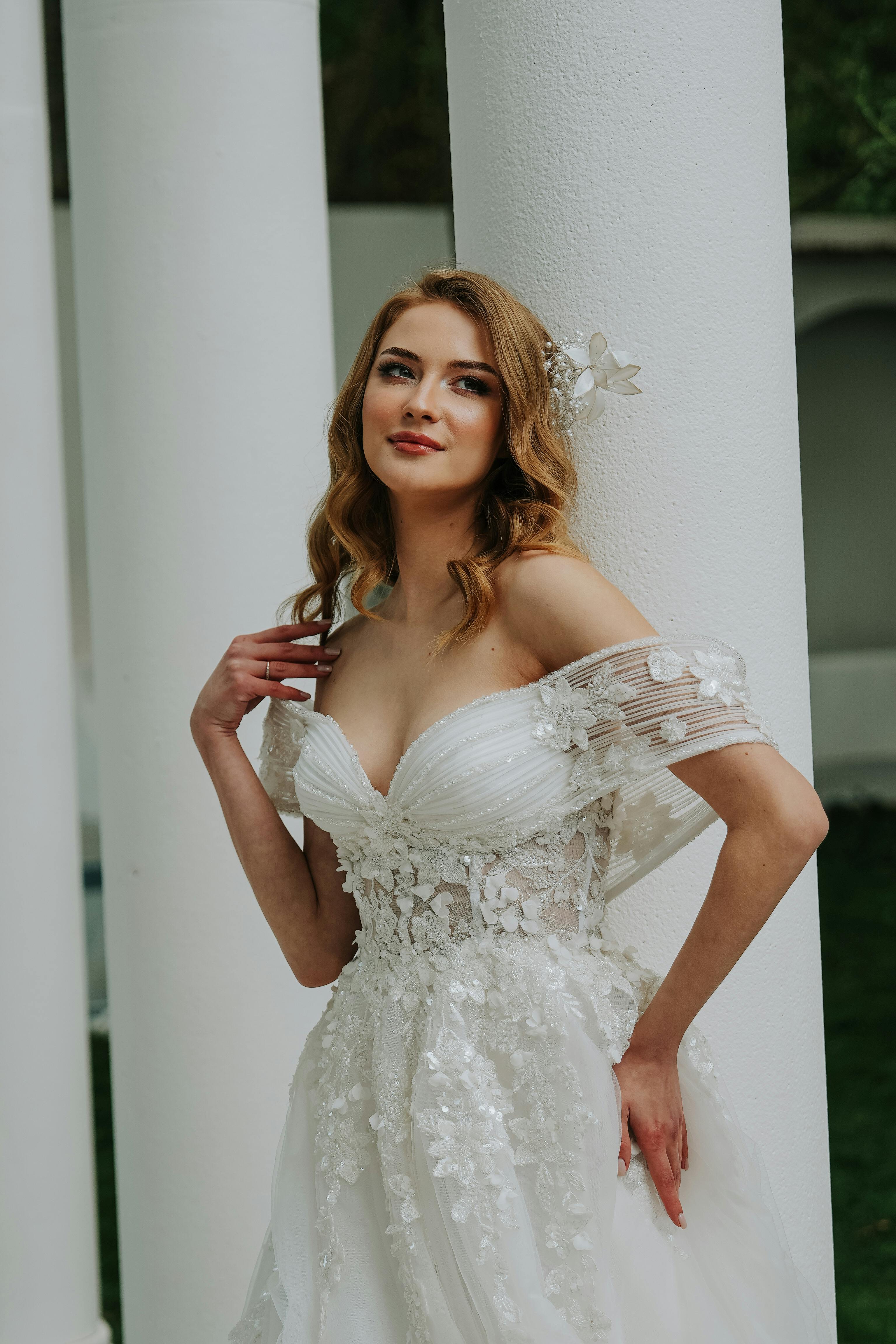 A beautiful bride in a white lace wedding dress poses gracefully against a column outdoors.