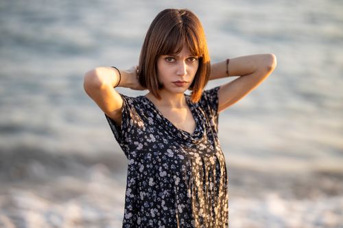 Free Young woman in floral dress poses by the sea during golden hour. Stock Photo