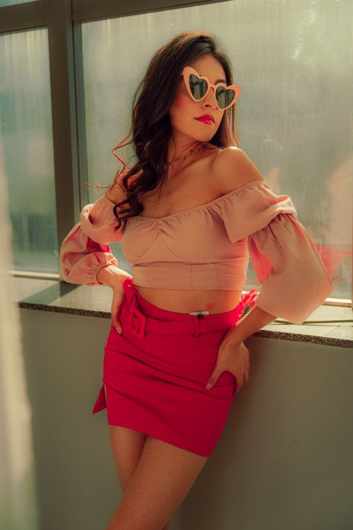 Free Elegantly posed woman in pink attire and sunglasses, basking in warm daylight by a window. Stock Photo