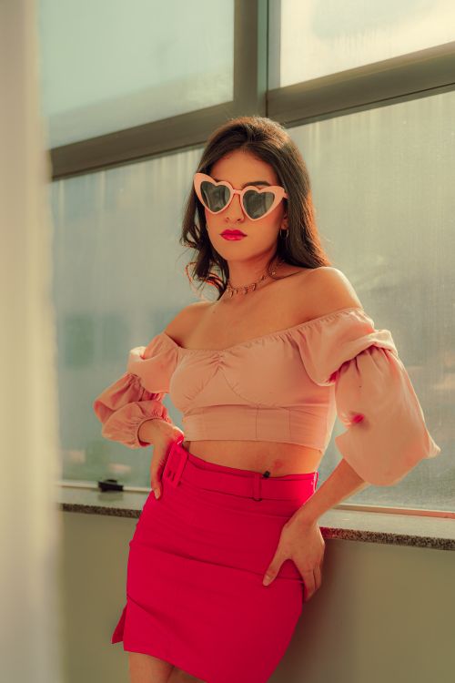Free Confident woman posing indoors wearing heart-shaped sunglasses, a pink blouse, and skirt. Stock Photo