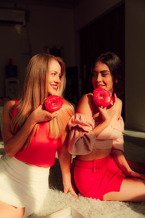 Free Two young women share a fun moment indoors, savoring vibrant red doughnuts in warm, soft lighting. Stock Photo