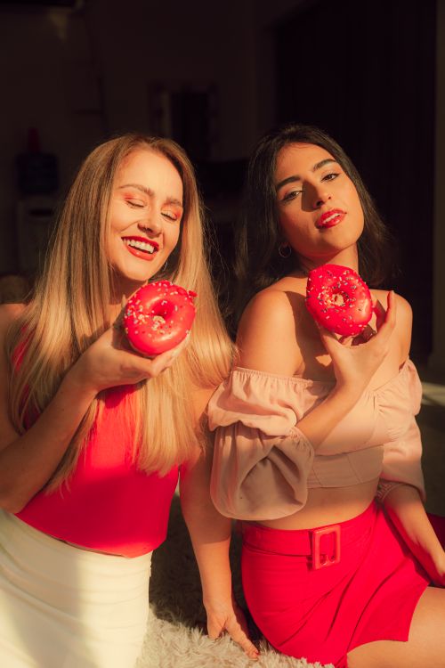 Free Two young women in bright attire enjoying donuts, basking in warm sunlight indoors. Stock Photo