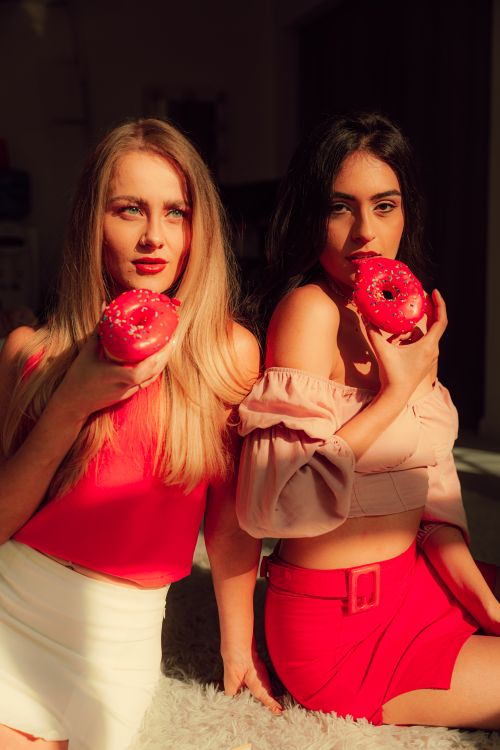 Free Two stylish women savoring pink donuts, bathed in warm sunlight. Fashion and indulgence. Stock Photo