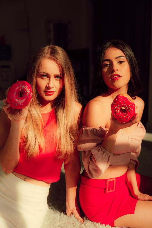 Free Two women pose in vibrant outfits, each holding a donut in warm light. Stock Photo