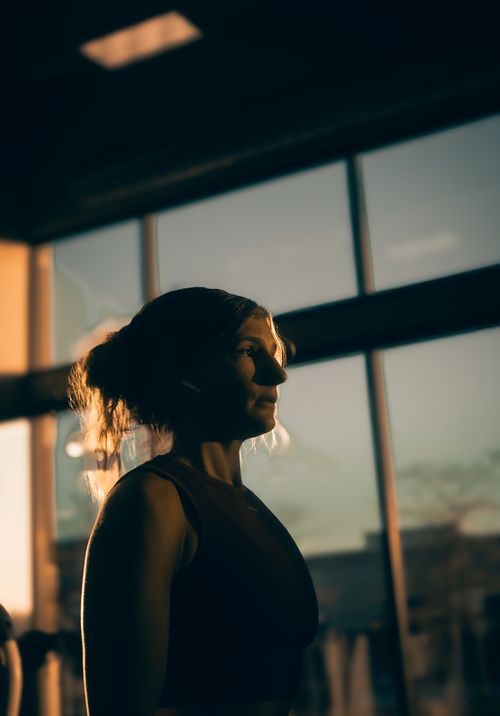 Free A woman stands thoughtfully in a gym, illuminated by the warm glow of the sunset. Stock Photo
