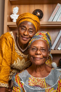 Two elderly African women wearing traditional attire, smiling indoors.