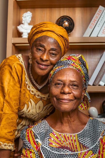Two elderly African women wearing traditional attire, smiling indoors.