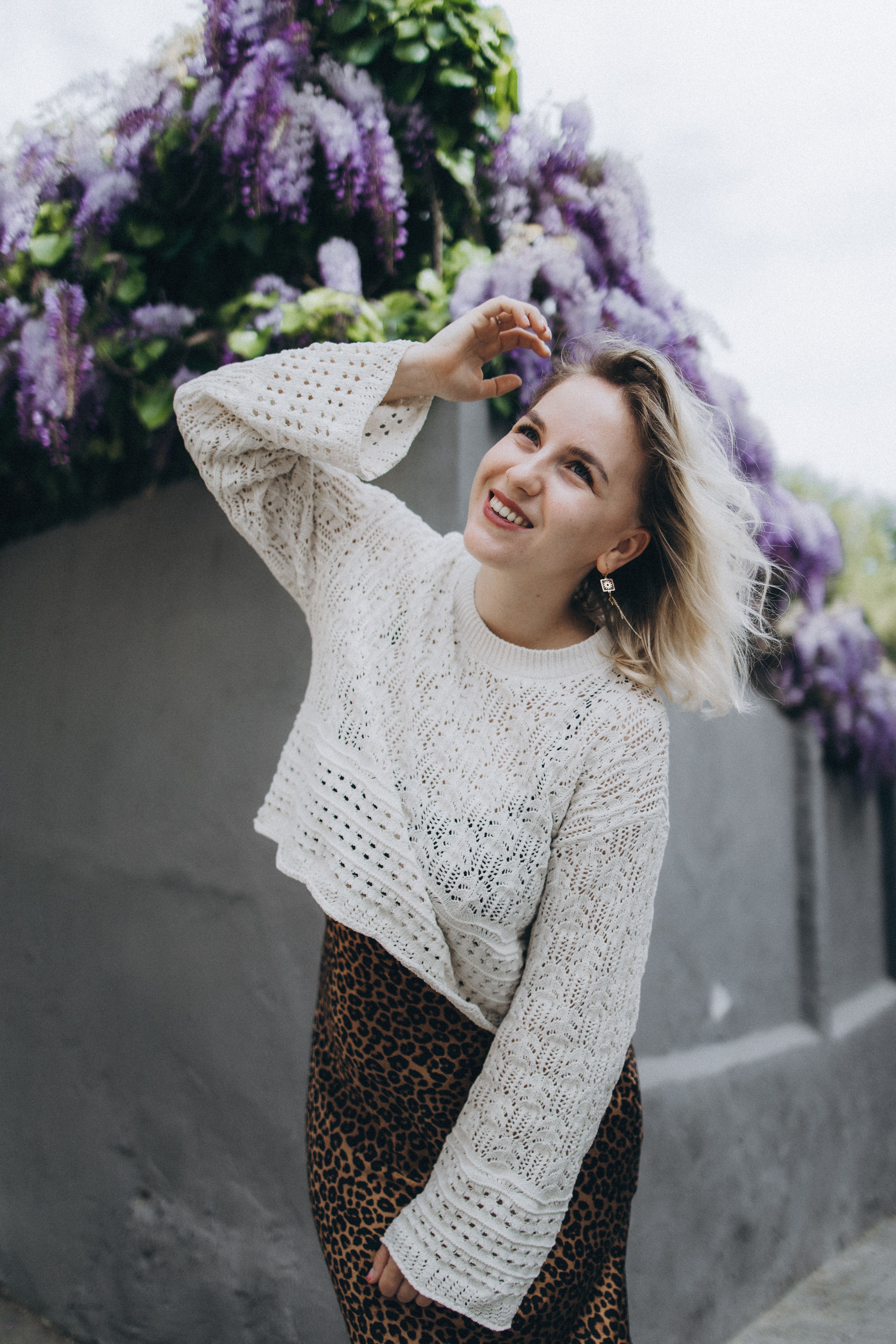 Joyful woman poses in a knitted sweater near vibrant purple flowers outdoors.