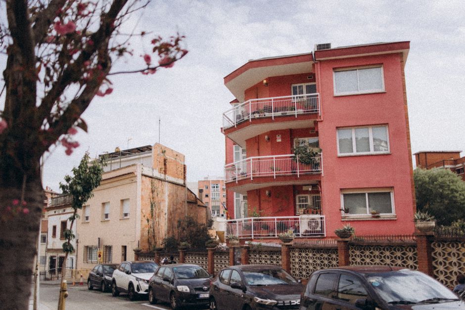Un edificio de apartamentos de color rosa vibrante en medio de una calle de la ciudad con autos estacionados y árboles en flor.
