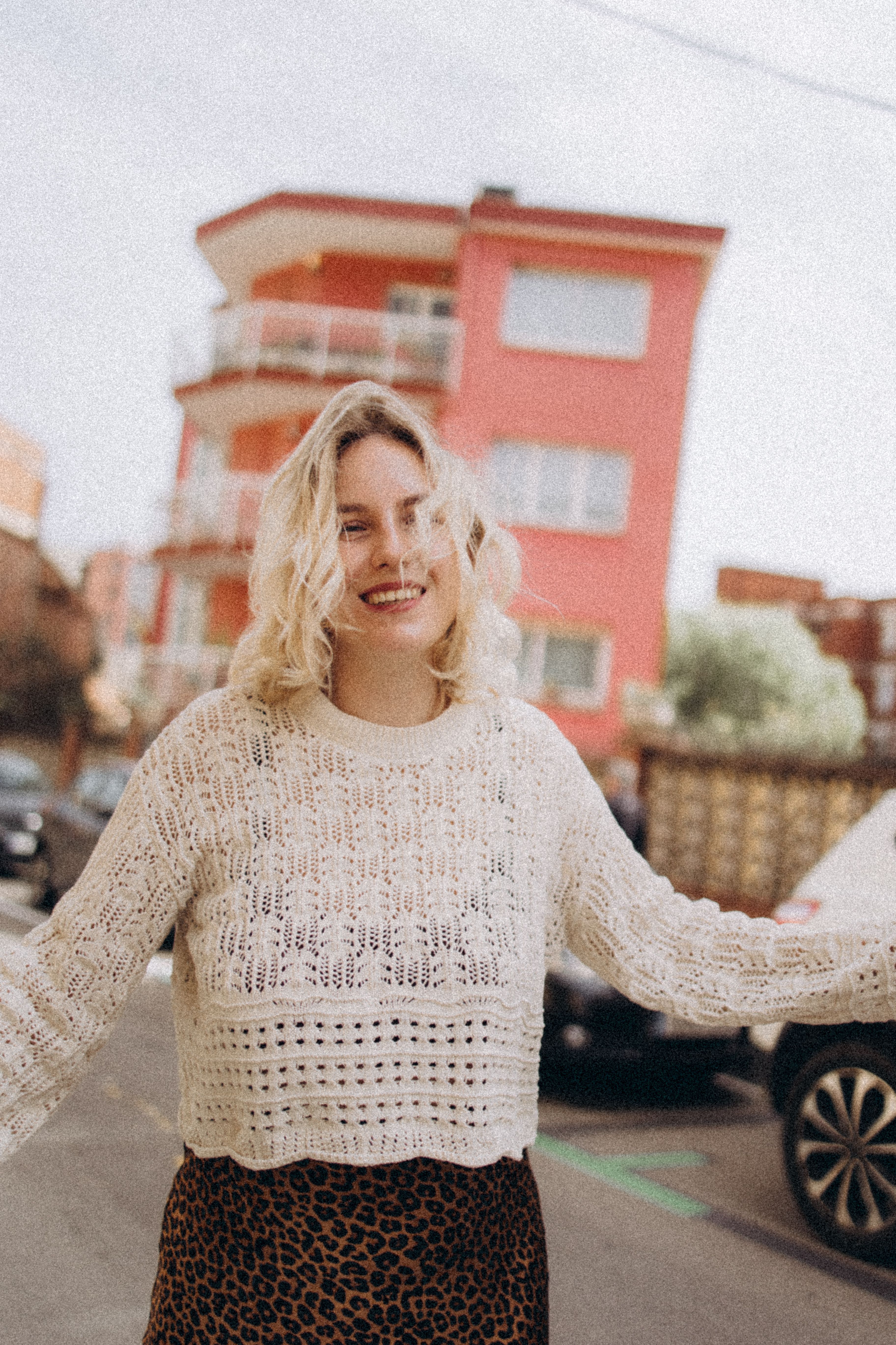 A cheerful woman in white lace sweater outdoors, urban background with a pink building.