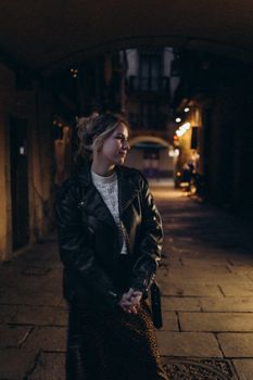 A young woman in a leather jacket poses thoughtfully in a dimly lit urban alleyway at night.