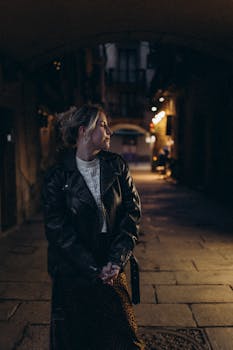 A woman in a leather jacket standing pensively in a dimly lit urban alley at night.