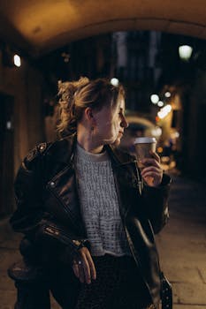 Fashionable woman in leather jacket enjoying coffee during nighttime stroll in city alley.