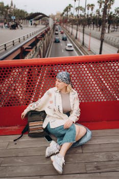 Stylish woman seated on bridge captures urban lifestyle, framed by red railing and modern architecture.