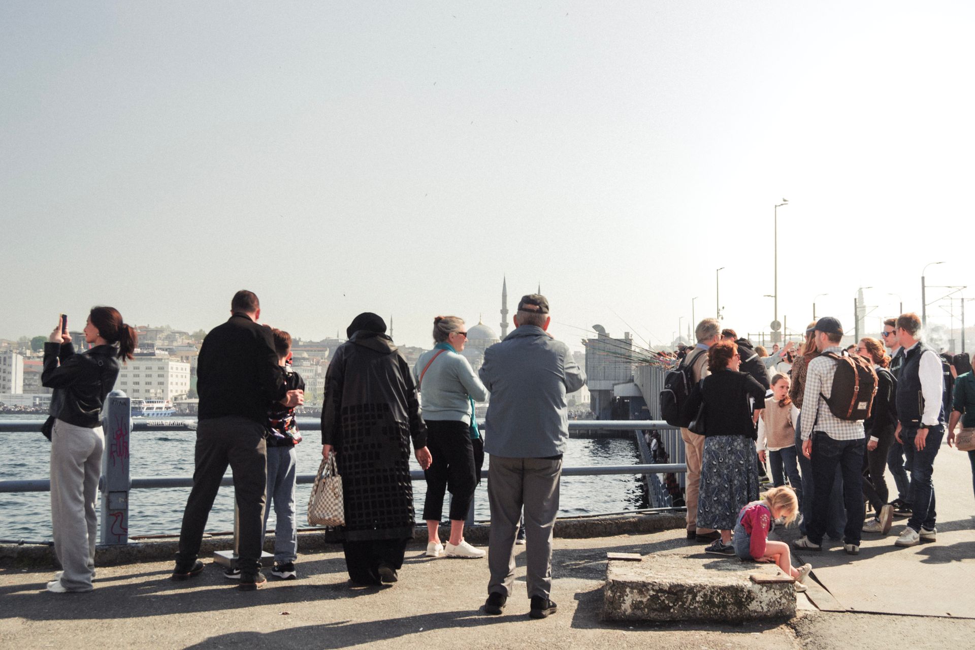 A diverse group of people gather on a riverside bridge enjoying the view on a sunny day.